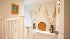 Un escalier avec une rampe blanche, un tableau encadré accroché au mur et deux vases contenant de hautes tiges de roseaux, placés sur une étagère.