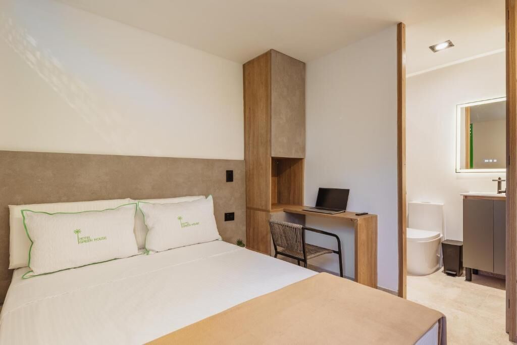 A hotel room with a bed, a desk with a laptop, a chair, a small trash bin, and a mirror in the bathroom.
