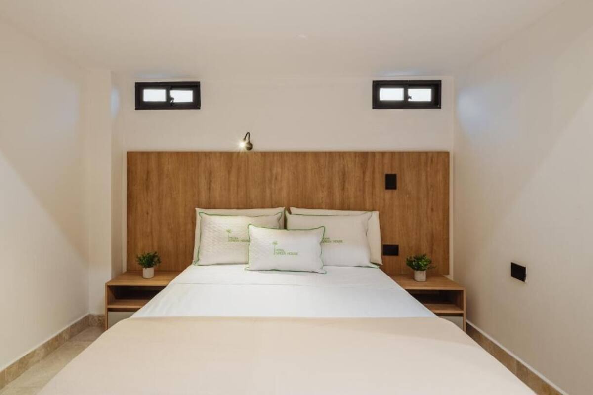 A bedroom with a wooden headboard, two bedside tables with plants, a bed with white bedding, and two small windows.
