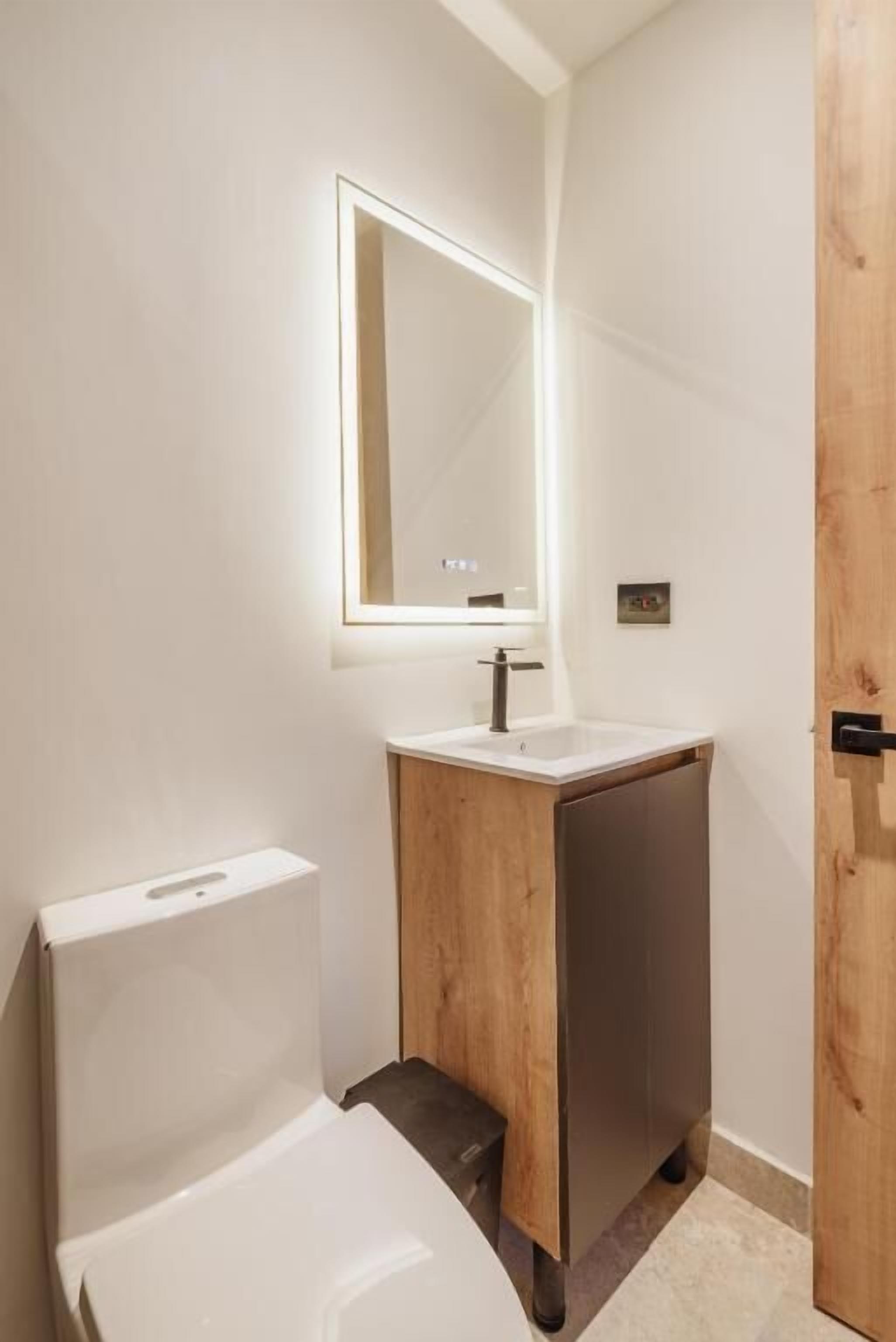 Un baño moderno con inodoro blanco, tocador de madera y espejo con luz superior.