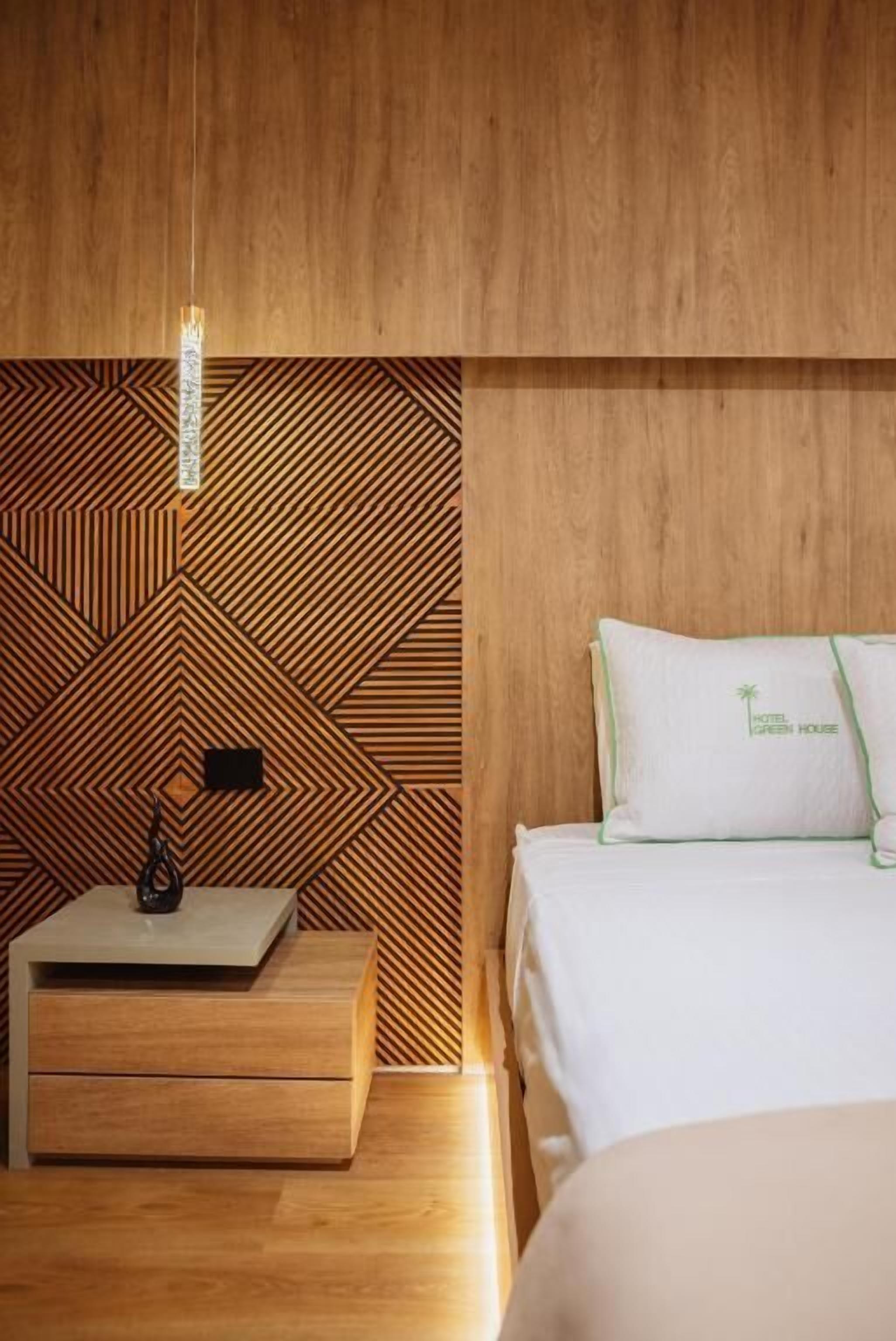 Une chambre d’hôtel avec un mur orné d’un motif géométrique, un lit avec un oreiller portant l’inscription « Hotel Green House », une table de chevet avec une lampe et une commode en bois.