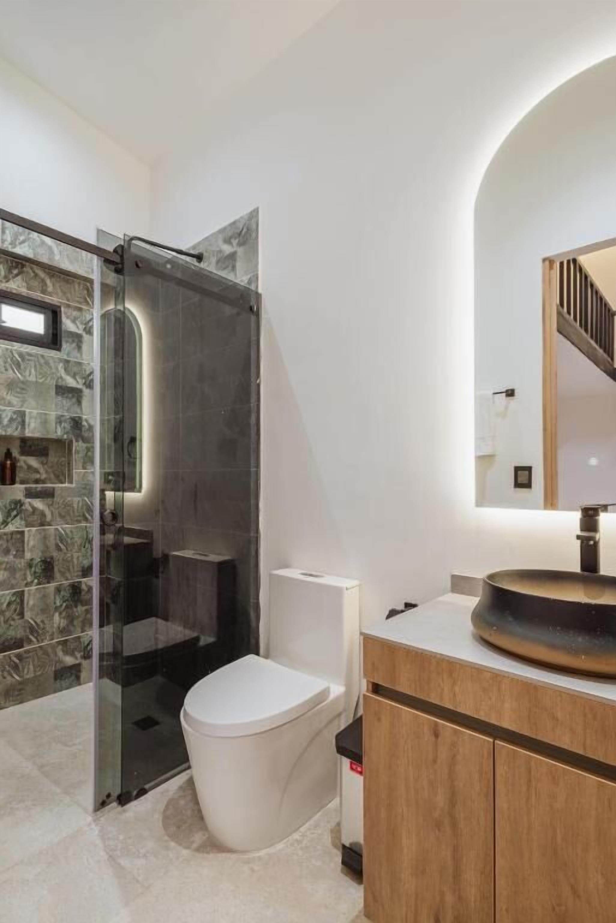 Un baño moderno con cabina de ducha de vidrio, inodoro blanco, tocador de madera y un espejo grande.