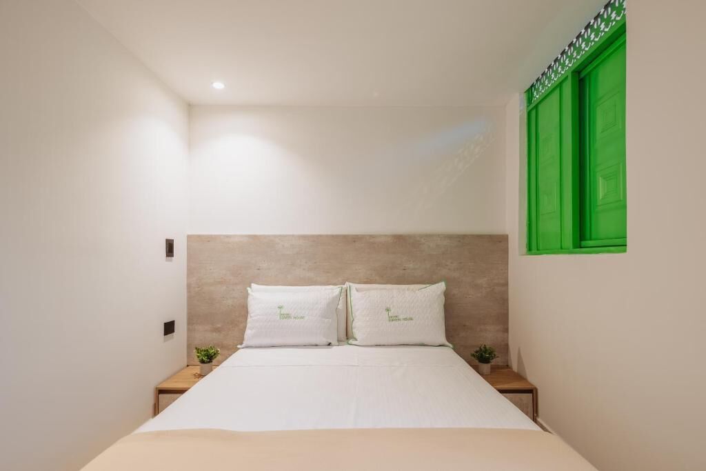 Un dormitorio con una cama, dos almohadas, una mesita de noche, una planta pequeña y un persiano verde.