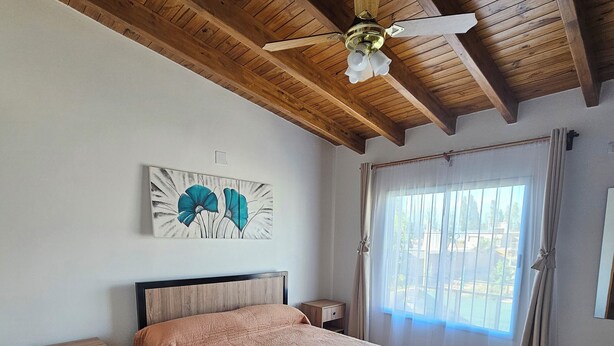 Een slaapkamer met een houten plafond, een bed met een beige dekbedovertrek, een nachtkastje, een aan de muur hangend schilderij en een raam met gordijnen.