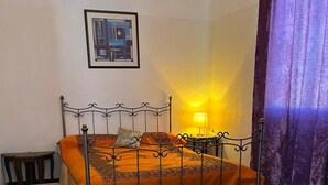 A single bed with an orange bedspread, a metal headboard, a small bedside table with a lamp, a checkered floor, and a purple curtain. - Rooftop del quinto (Fès)