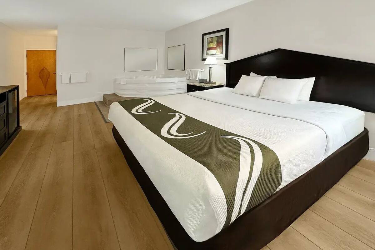 A hotel room with a large bed, a wooden floor, a bathroom with a bathtub, and a door leading to another room.