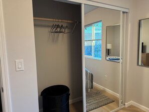 Una habitación con una puerta corredera que da a un baño, un cesto de basura negro, un perchero con ganchos, una ventana con persianas y un espejo en la pared.