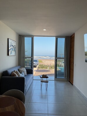 Een moderne woonkamer met een grote schuifglazen deur die uitkomt op een balkon met uitzicht op een strand.