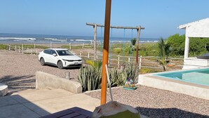 Ein Holztisch mit einer Kokosnuss auf einem Teller, einem Sonnenschirm und einem weißen Auto, das auf einem Schotterweg mit Blick auf das Meer und einen Pool geparkt ist.