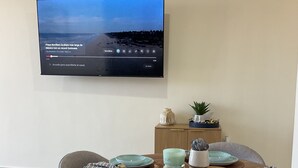 Un coin repas avec une table ronde en bois, dressée pour quatre personnes, entourée de chaises à revêtement gris. Un téléviseur à écran plat, fixé au mur, diffuse une chaîne d’information.