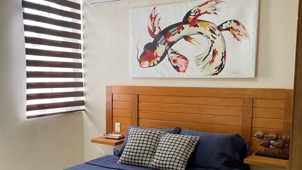 Une chambre à coucher comprenant un lit, une tête de lit en bois, une table de chevet, un climatiseur mural et une fenêtre avec des stores.