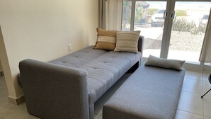 Ein kleines, graues Schlafsofa mit einer Bank, einer Steckdose und Meerblick durch ein großes Fenster.