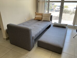 A compact, grey sofa bed with a bench, a power outlet, and a view of a beach through a large window. - Fabulous oceanfront 2-bedroom condo with AC in Playa Novillero (Playa Novillero)
