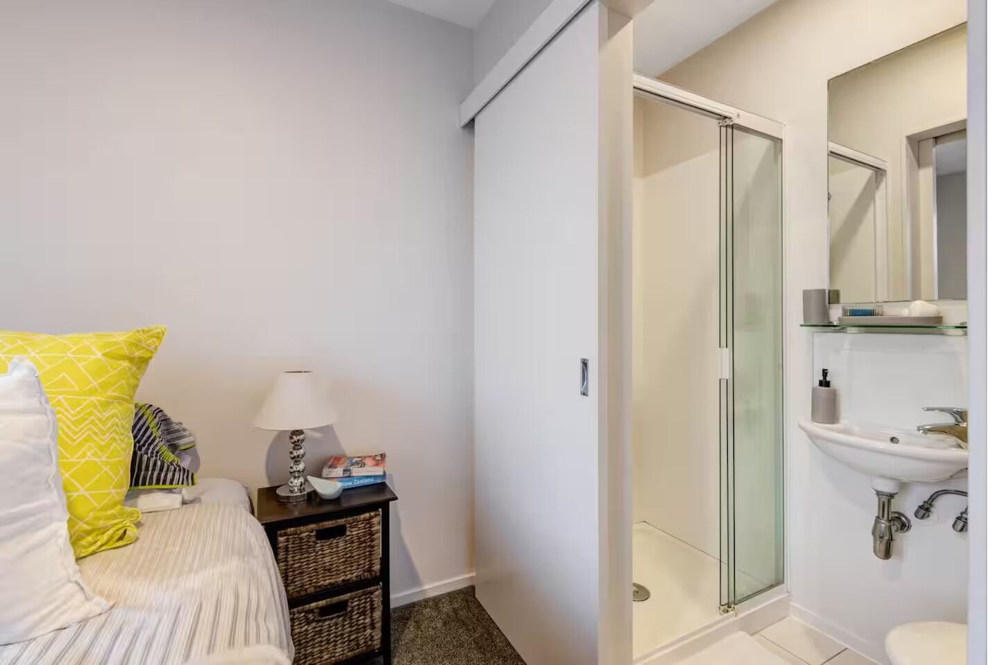 Une chambre à coucher avec un lit, une table de chevet avec une lampe et des livres, une petite commode et une cabine de douche en verre.