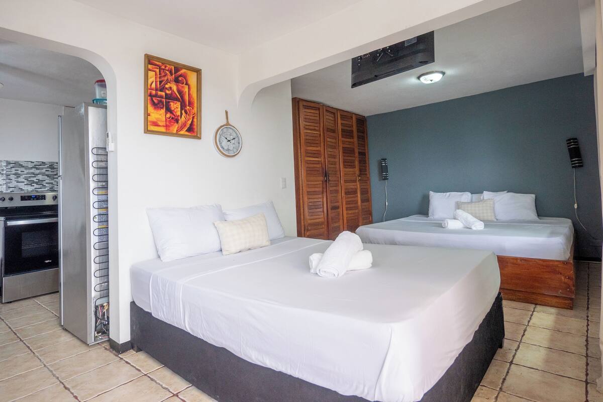 A hotel room with two beds, a kitchenette, a clock, and a painting on the wall.