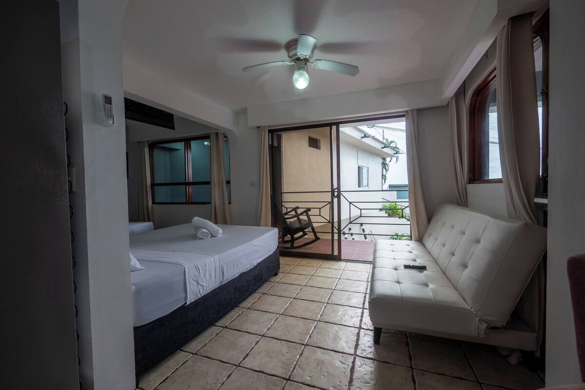 A hotel room with a bed, a sofa, a ceiling fan, and a view of the outside.
