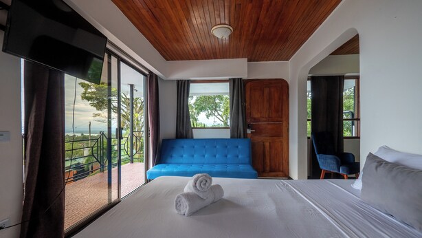 A hotel room with a bed, a blue sofa, a wooden door, and a view of greenery. - Hotel Manuel antonio beracha (Quepos)