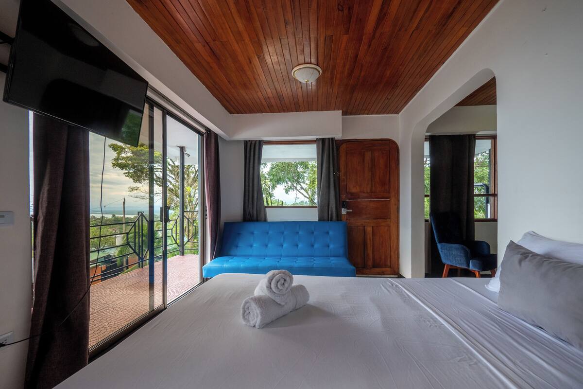 A hotel room with a bed, a blue sofa, a wooden door, and a view of greenery.