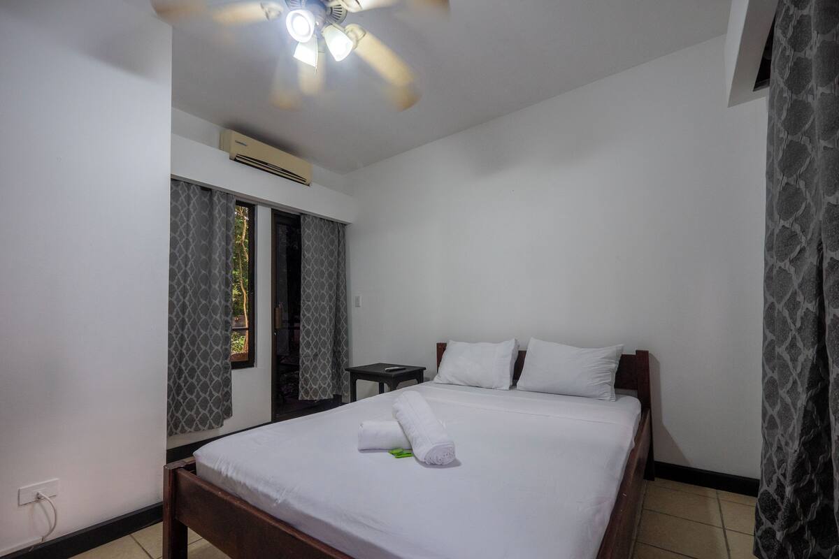 A hotel room with a bed, a ceiling fan, an air conditioning unit, a window with curtains, and a patterned curtain on the right.