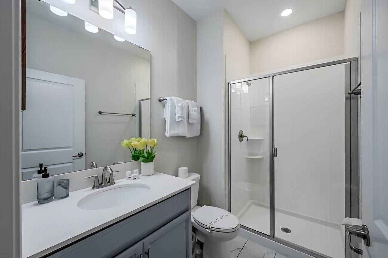 A modern bathroom with a large mirror, a sink with a vase of flowers, a toilet, and a glass-enclosed shower.