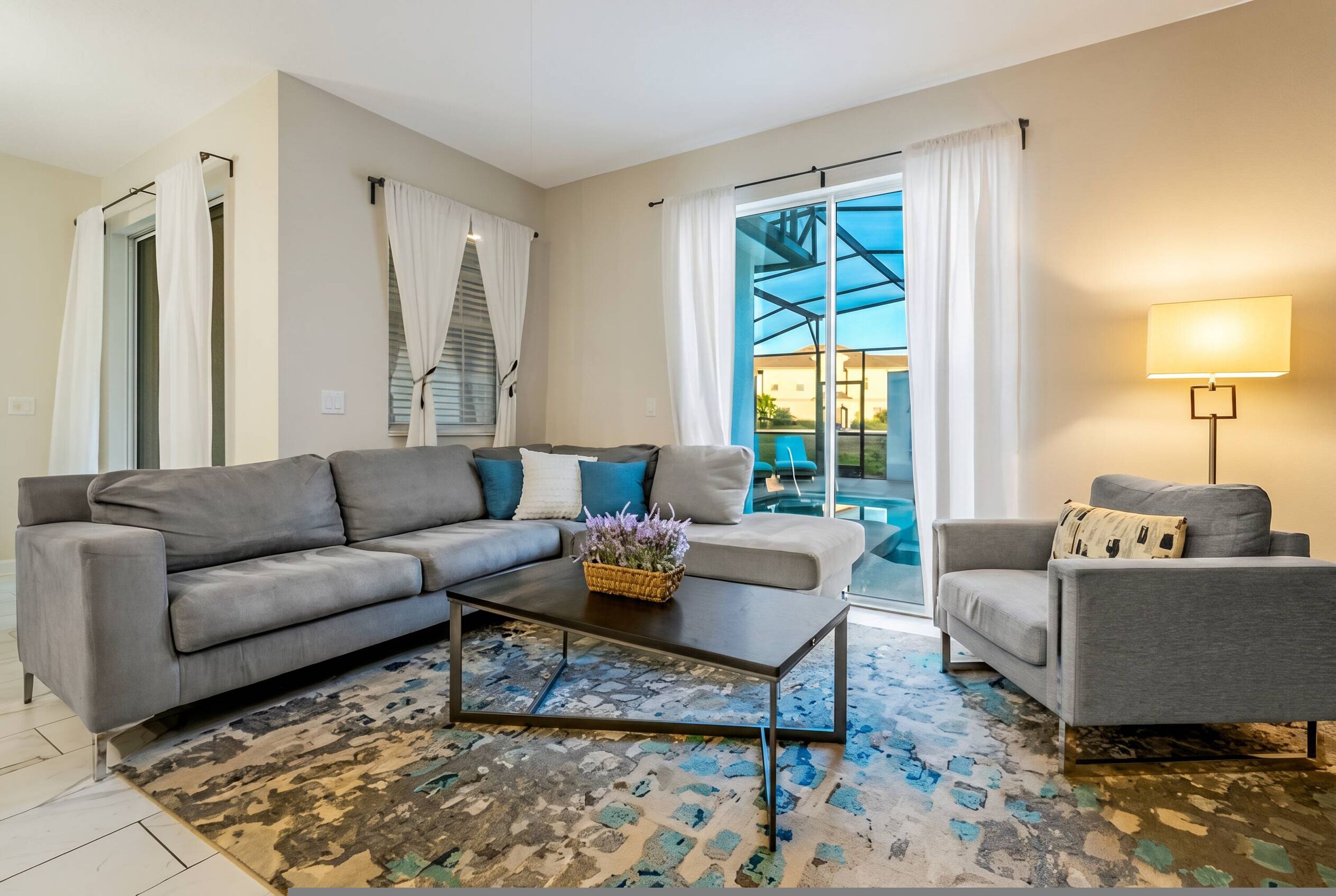 A living room with a grey sofa, a smaller armchair, a coffee table, and a lamp. There is a glass door leading to an outdoor area.