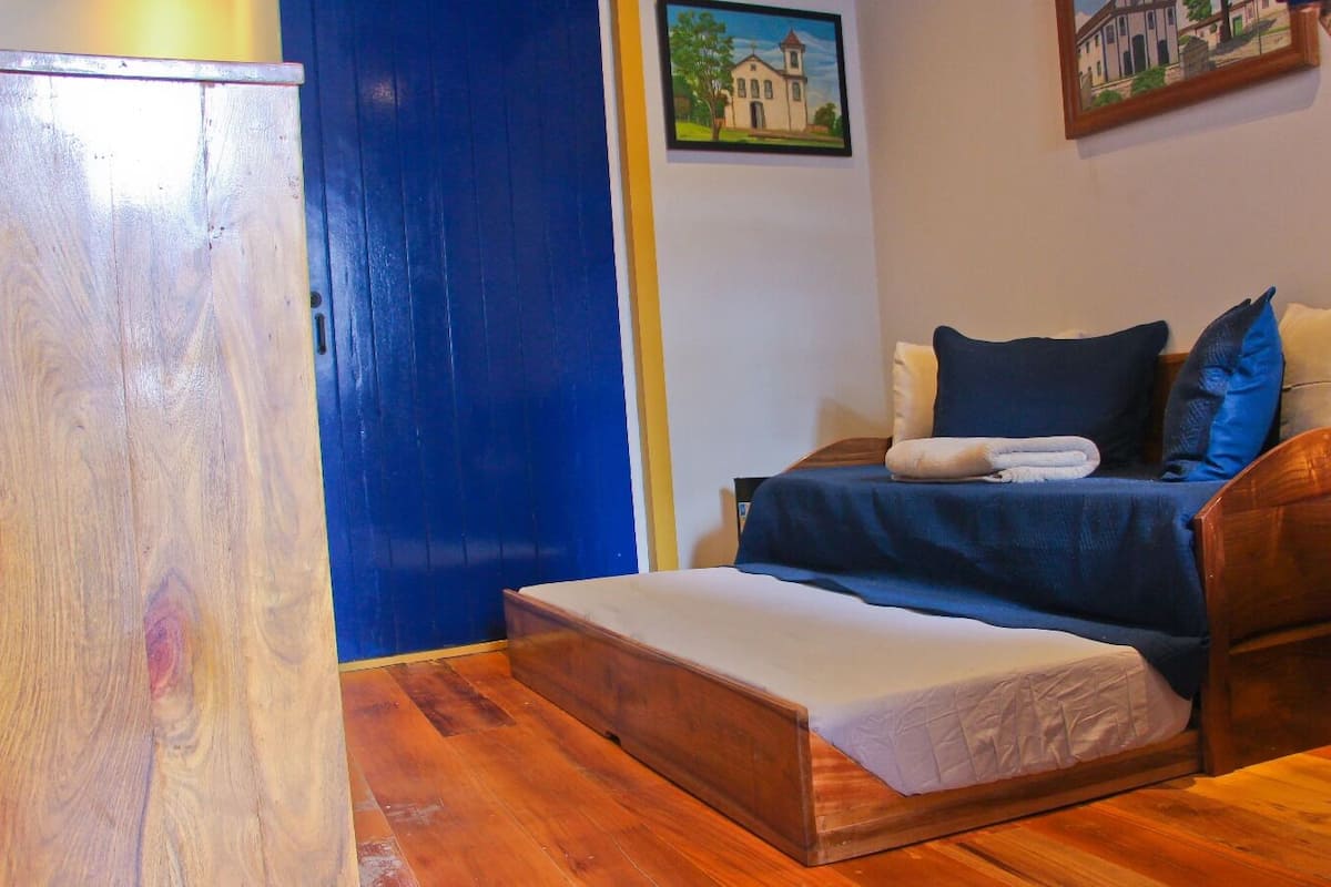 A small, cozy room with a wooden floor, a bed with a blue blanket and pillows, and a small bench.