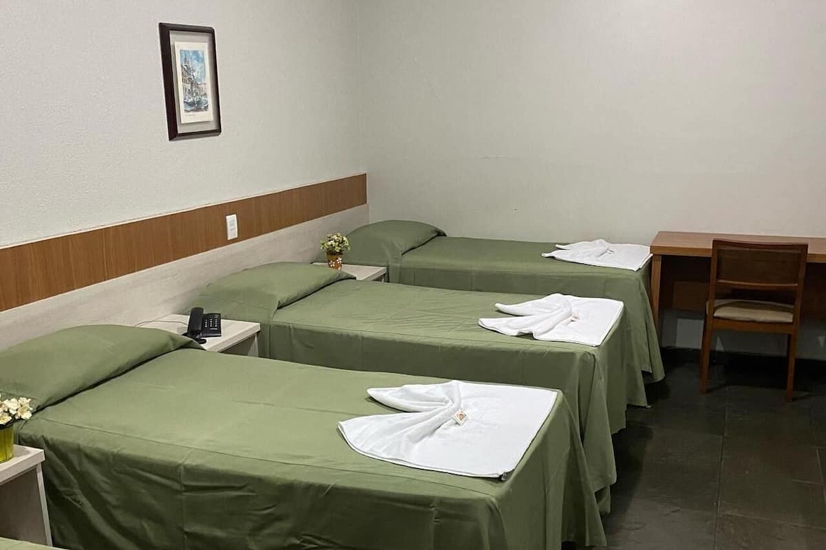 A hotel room with four single beds, each with a green bedspread and white towels. There is a wooden desk and a chair to the right, and a small table with a vase of flowers on the left.