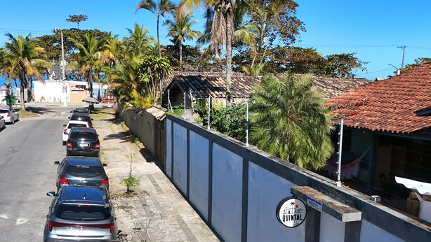 A row of parked cars along a street, a building with a sign reading 'QUINTAL HOSTEL,' and a fenced area with palm trees. - HOSTEL QUINTAL GUEST - Guarujá (Guarujá)