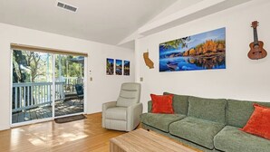 A spacious living room with a green sofa, a grey armchair, and a wooden coffee table. A large window offers a view of a deck and trees. - Yosemite Terrace View Retreat (Mariposa)