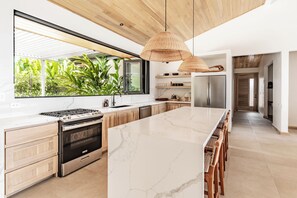A modern kitchen with a large island, stainless steel appliances, and a view of greenery outside.