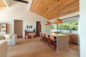 A modern kitchen with a central island, wooden ceiling, and a view of greenery outside.