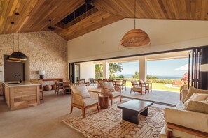 A spacious living area with a stone wall, wooden ceiling, and large windows offering a view of the outdoors. - Casa Palmera Azul (Osa)