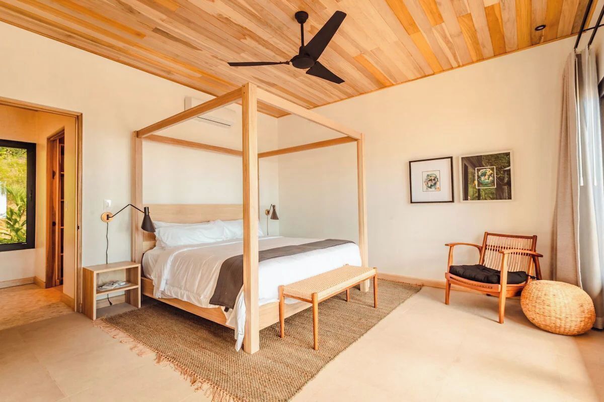 Un dormitorio con una cama con dosel, un ventilador de techo de madera, una silla y una cesta tejida.