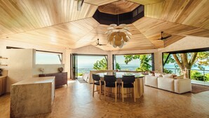 Un interior espacioso y moderno con techo de madera, zona de comedor central y ventanas amplias con vistas al océano.