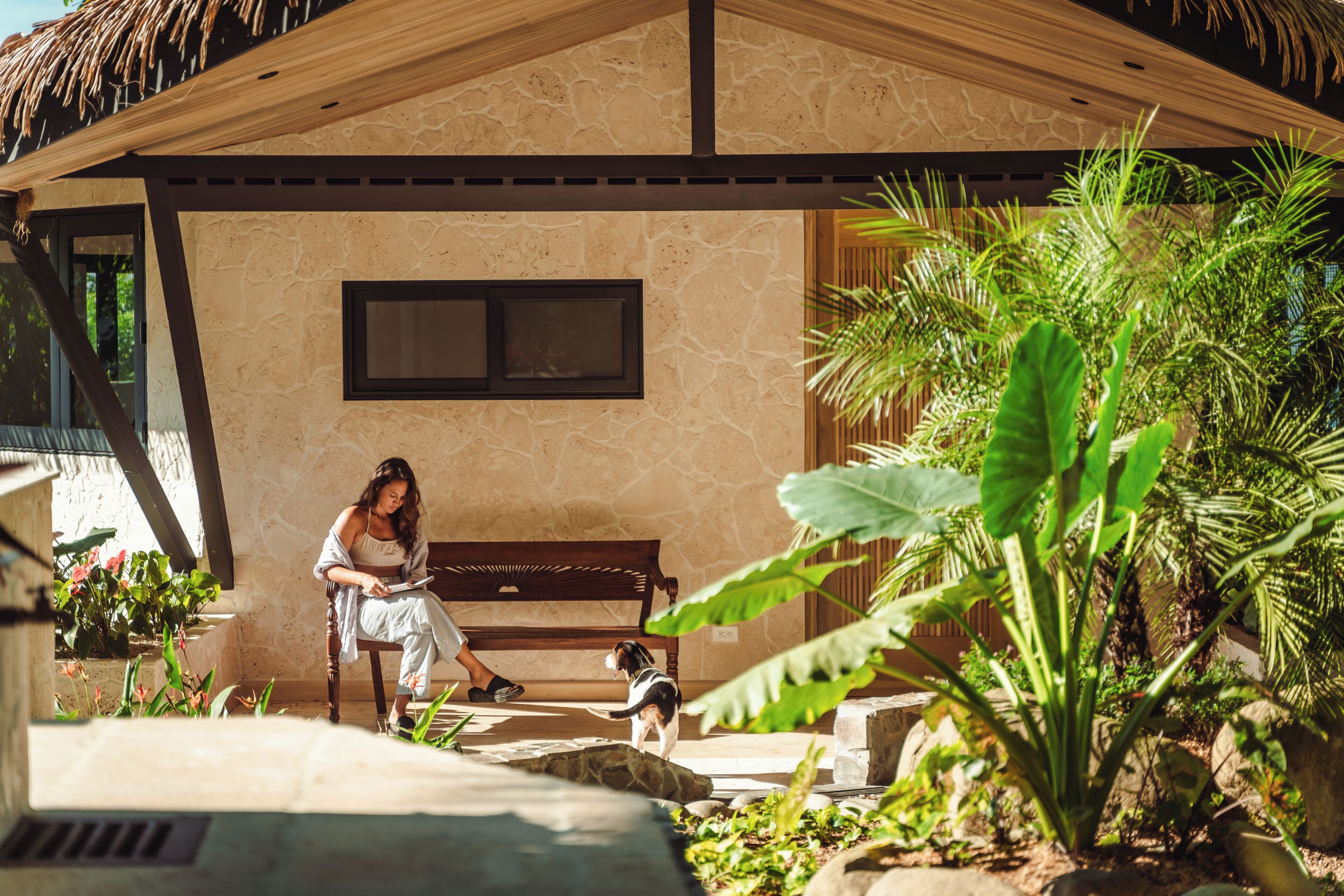 A covered outdoor seating area with a stone wall, a bench, and lush greenery.