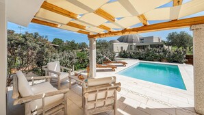 A poolside area with wooden chairs and a table, a covered pergola, and a view of a modern building in the background. - Nice home in San Teodoro (San Teodoro)