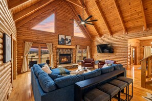 Interior de una cabaña de troncos con un sofá grande, chimenea, televisión y una vista panorámica a través de las ventanas.