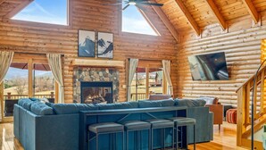 A log cabin interior with a large sofa, a fireplace, a TV mounted on the wall, and a wooden ceiling. - Grand Mountain Lodge, Pickleball, Barrel Sauna, Hot Tub, Game Room, Arcade (Luray)