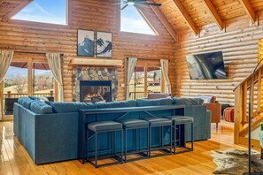 A log cabin interior with a large sofa, a fireplace, a TV mounted on the wall, and a wooden ceiling.