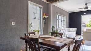 A dining area with a wooden table and chairs, a white sofa, and a window with a view of the outside. - Casa em Buzios com Piscina perto da Praia (Armação dos Búzios)
