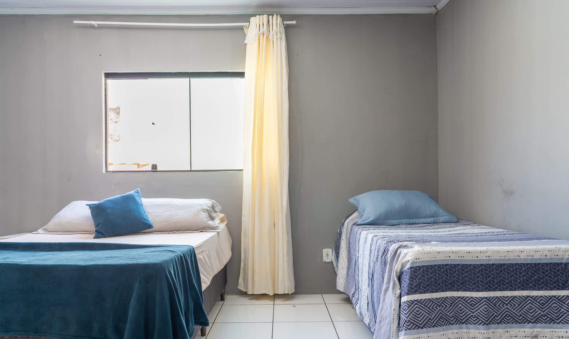 A room with two beds, a window with a curtain, and a wall with a visible electrical outlet.