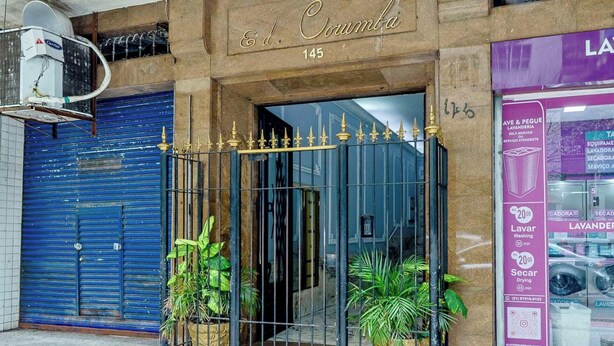 A building entrance with a blue shutter door, a metal gate with decorative elements, and a sign above the door. - Apto no Rio a 200m da Praia de Copacabana (Rio de Janeiro)