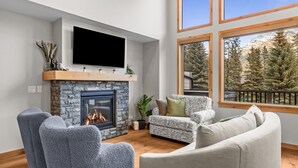 A modern living room with a stone fireplace, a mounted TV, and large windows offering a mountain view. - Luxury Modern Mountain Lodge in Spring Creek, Canmore (Canmore)