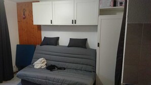 Quarto com cama, duas almofadas, um cobertor e uma mochila. Há um painel de madeira na parede e um armário branco acima da cama.