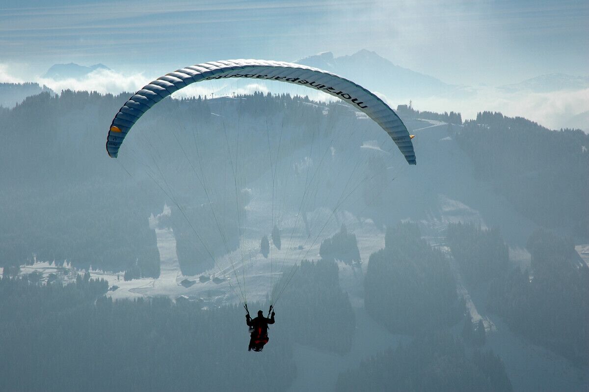 Eine Person gleitet mit einem Paragleiter über eine gebirgige Landschaft unter blauem Himmel.