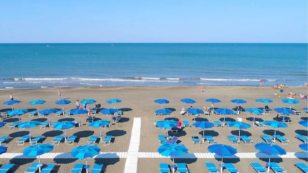 Ein Strand mit Reihen blauer Sonnenschirme und Liegestühle, ein klarer Himmel und das Meer im Hintergrund.