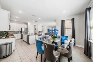 A modern kitchen with white cabinets, a stainless steel refrigerator, and a dining area with a glass table and blue chairs.