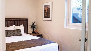 Ein Schlafzimmer mit Bett, Nachttisch, einer Blumenvase, einem gerahmten Bild an der Wand und einem Fenster mit Blick auf eine Treppe.