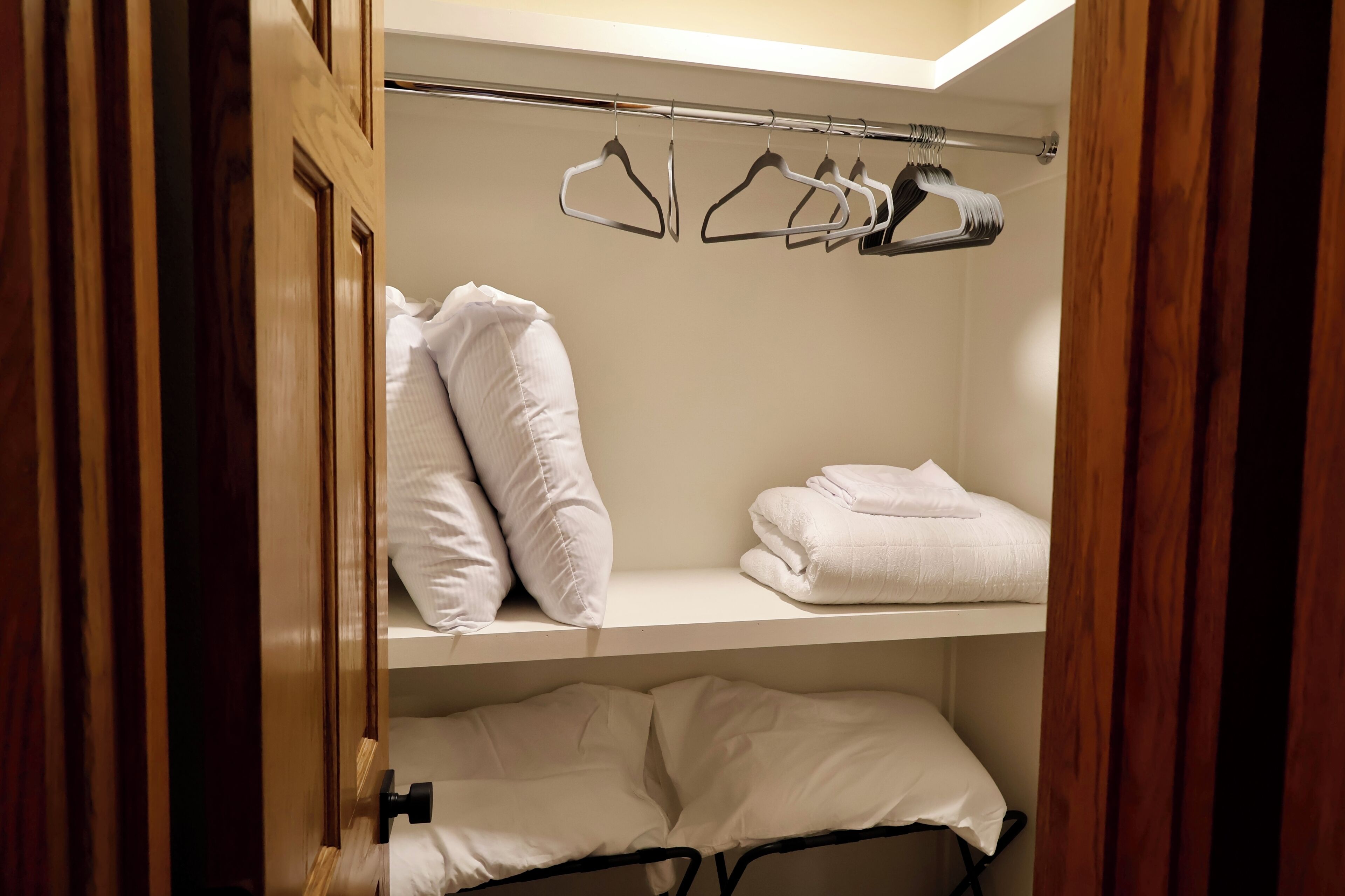 A hotel room with a wooden wardrobe, a bed with white bedding, and a shelf with folded towels and hangers for clothes.