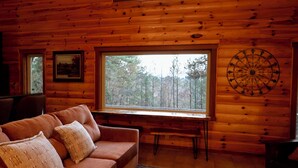 A log cabin interior with a wooden ceiling, a large window, a sofa with cushions, a framed picture, and a decorative metal piece on the wall. - Secluded Mountain Cabin near Scenic Lakes & Trails (Hot Springs)